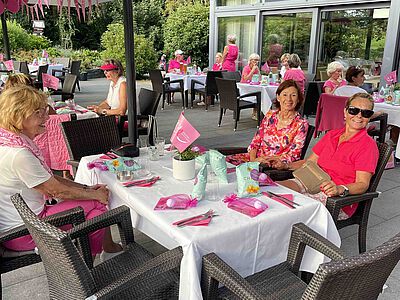 Damen sitzen an einem Tisch, weitere Damen im Hintergrund unter Sonnenschirmen.
